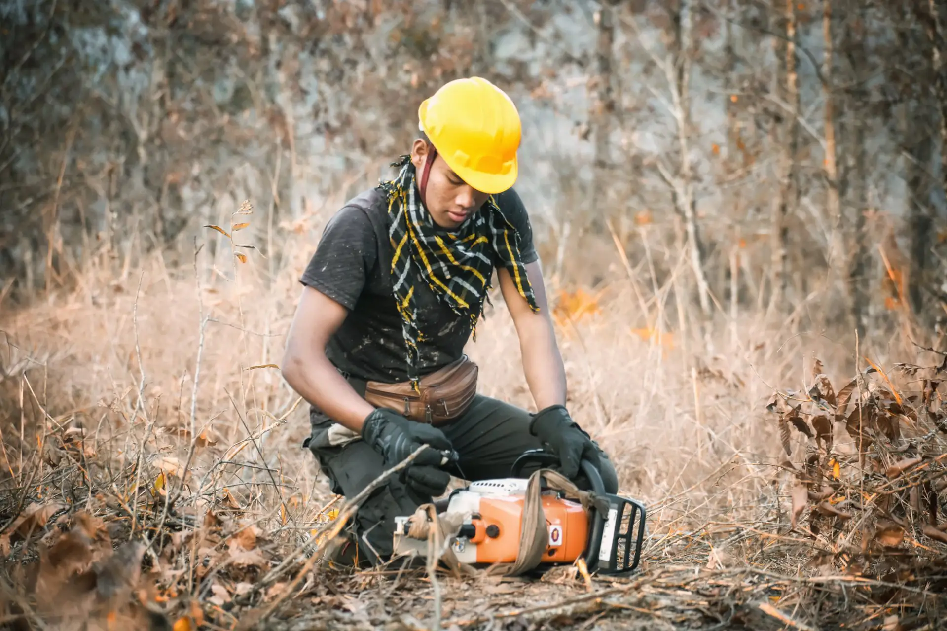 Worker in safety gear checking a chainsaw in a wooded clearing during brush removal