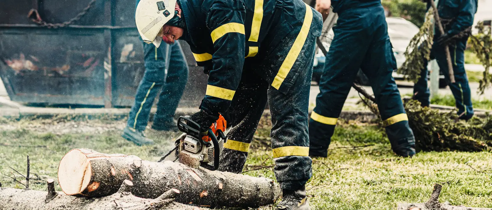 Crew in high-visibility gear bucking a downed trunk into sections with chainsaws after a storm